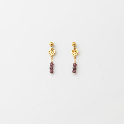 A pair of gold dangle earrings with a circular filigree disc, a small gold bead, and three dangling faceted dark red beads, shown on a plain white background.