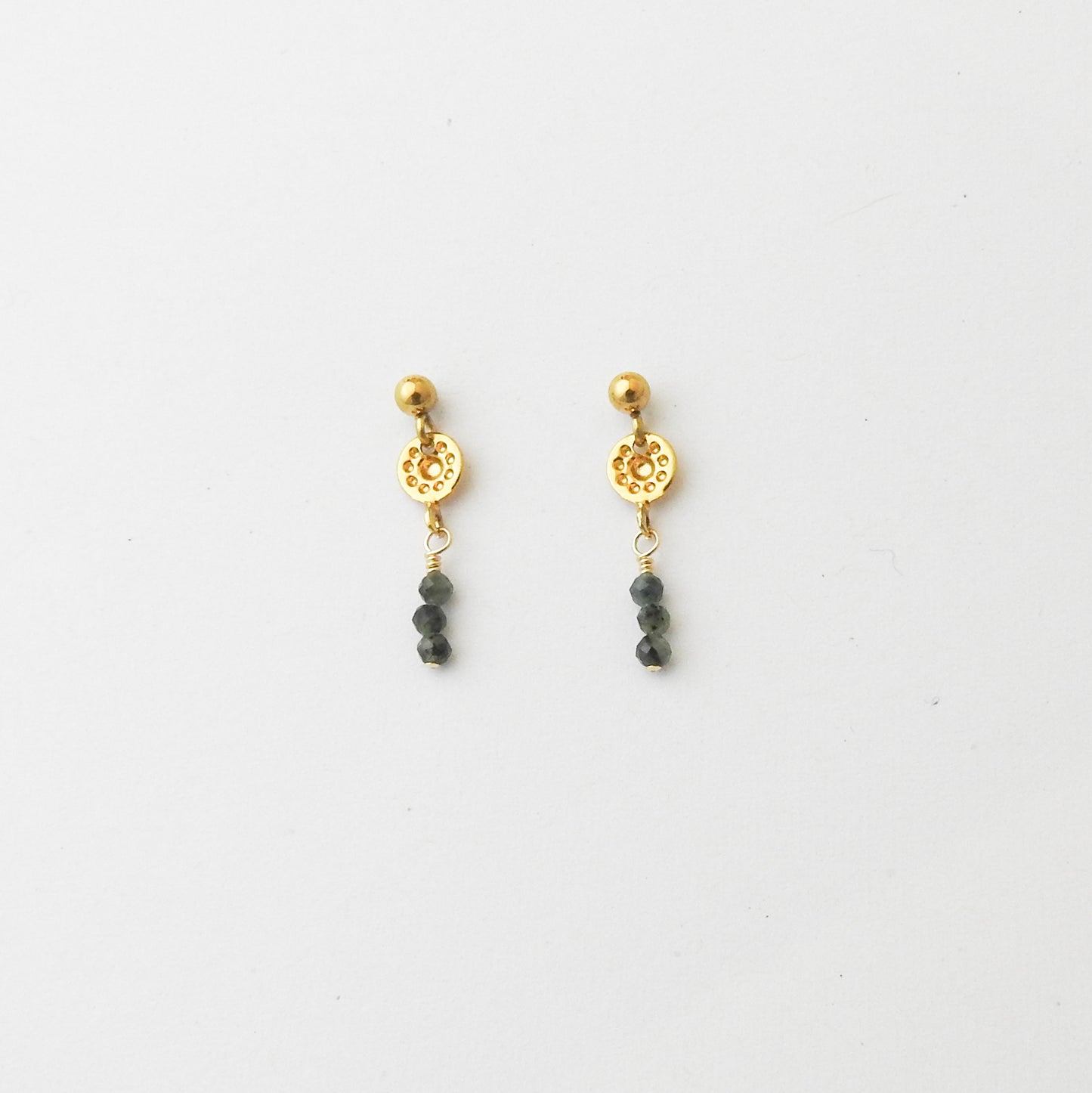 A pair of gold dangle earrings with a textured circular disc, a small gold bead, and three dark gray beads hanging below, displayed on a clean white background.