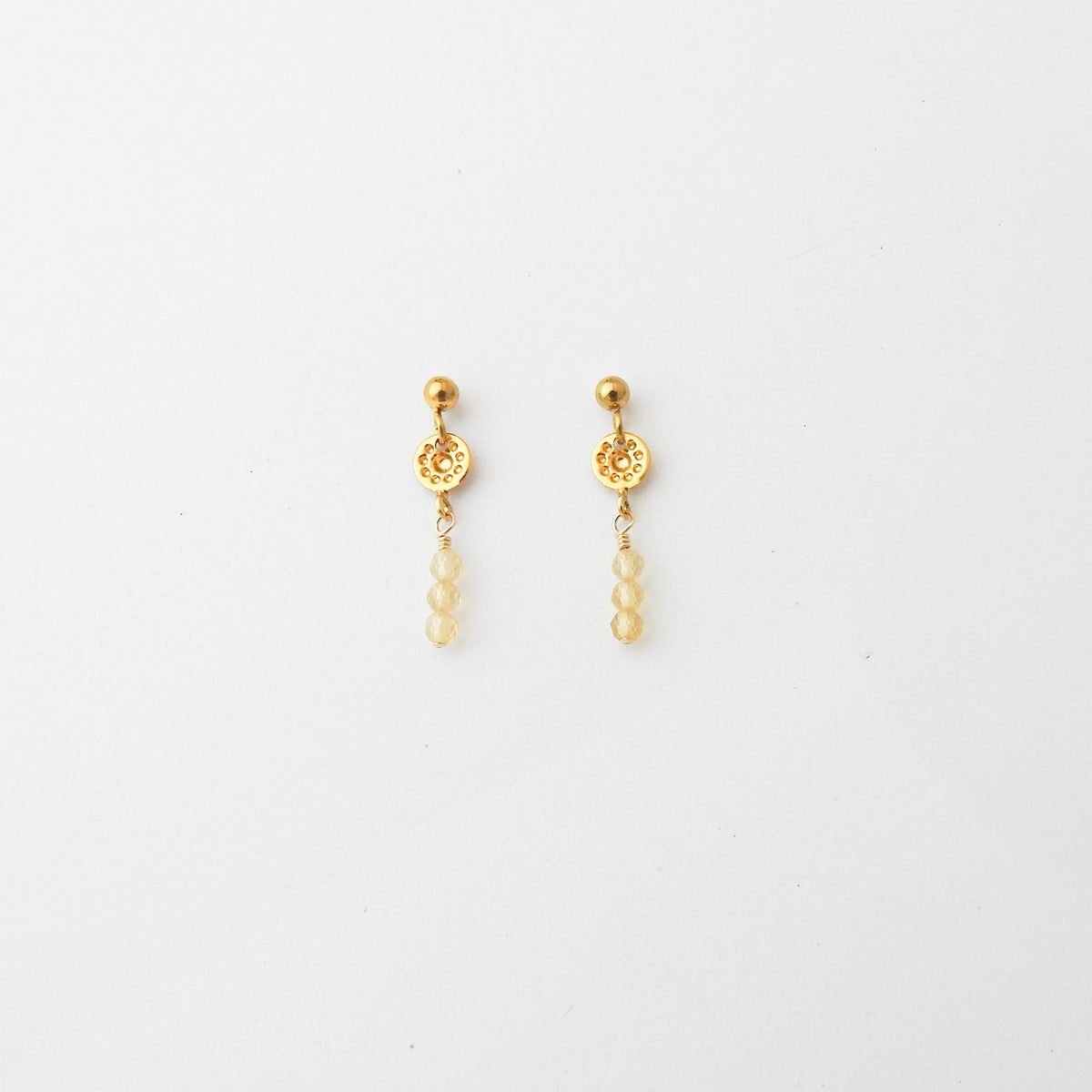 A pair of gold dangle earrings with a textured circular disc, a small gold bead, and three pale yellow beads hanging below, shown on a plain white background.