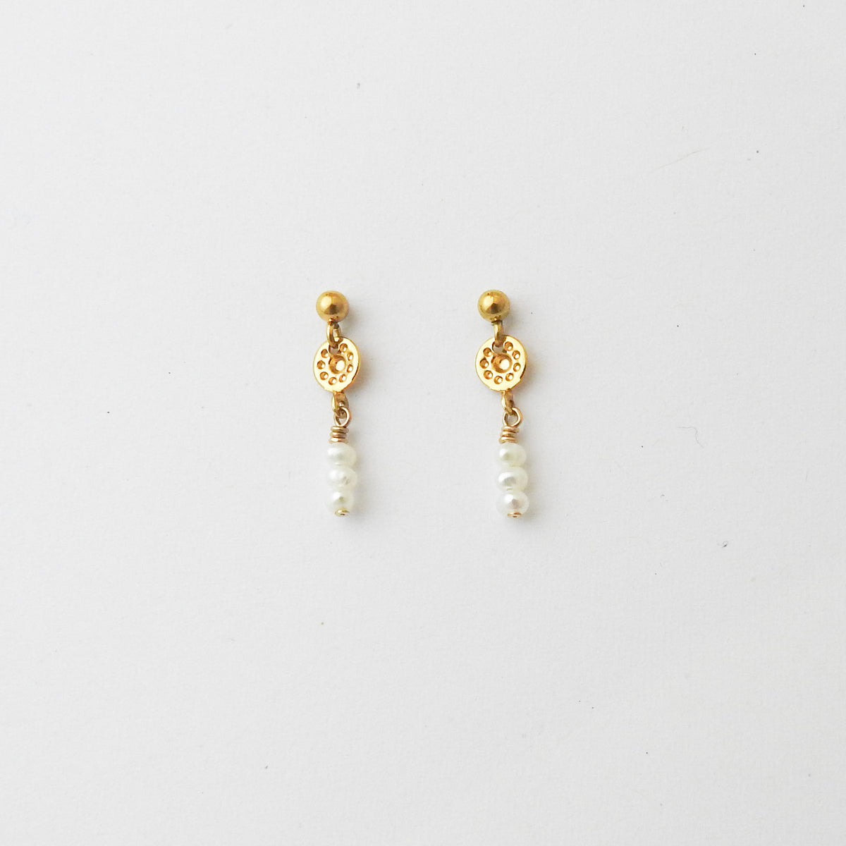 A pair of gold dangle earrings with a small round gold bead, an ornate circular gold disc, and three small white pearl beads hanging below, shown on a plain light gray background.