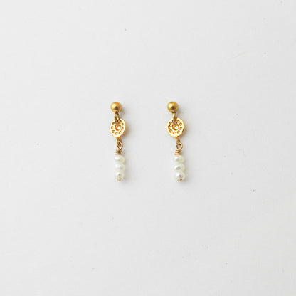 A pair of gold dangle earrings with a small round gold bead, an ornate circular gold disc, and three small white pearl beads hanging below, shown on a plain light gray background.