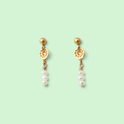 A pair of gold dangle earrings with circular filigree details, small gold beads, and three white pearl drops, set against a solid pale mint green background.
