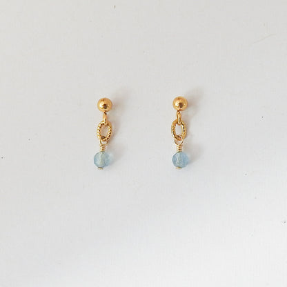 A pair of small gold dangle earrings with a faceted light blue bead at the bottom, displayed on a plain white background.