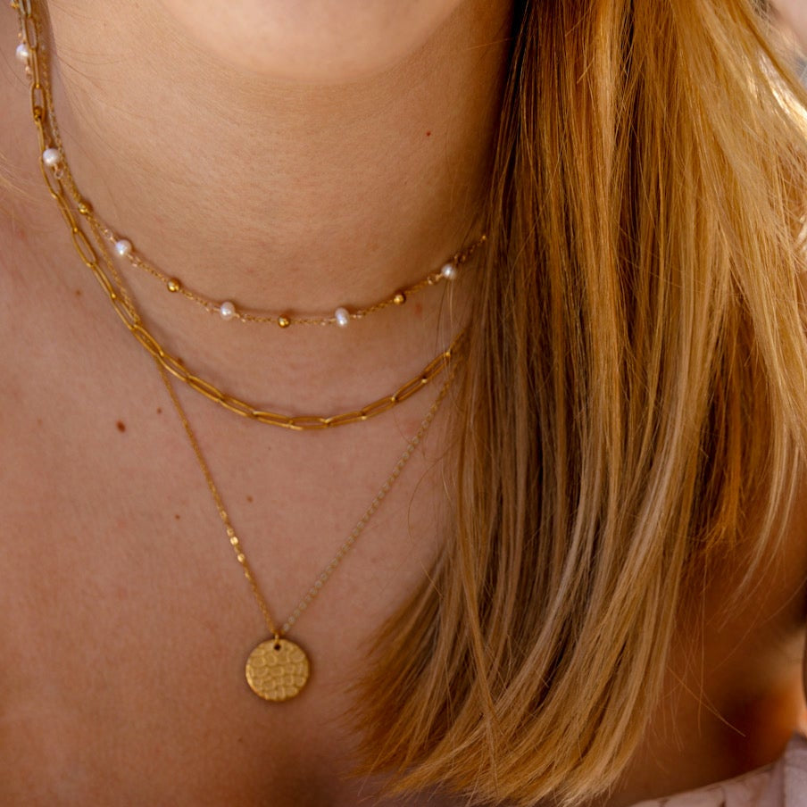 A close-up lifestyle shot of a woman wearing layered gold necklaces, bracelets, and a delicate chain earring, paired with a light pink ruffled off-the-shoulder top. A coffee cup is visible in the foreground on a gray stone tabletop.