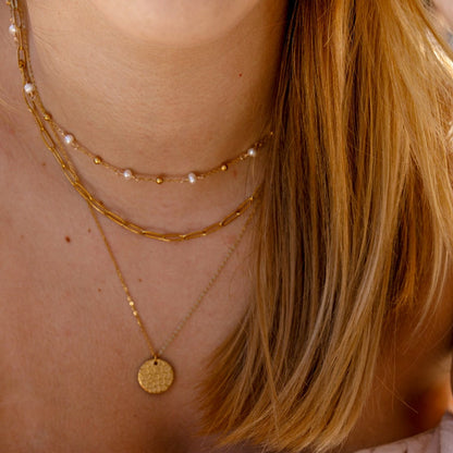 A close-up lifestyle shot of a woman wearing layered gold necklaces, bracelets, and a delicate chain earring, paired with a light pink ruffled off-the-shoulder top. A coffee cup is visible in the foreground on a gray stone tabletop.