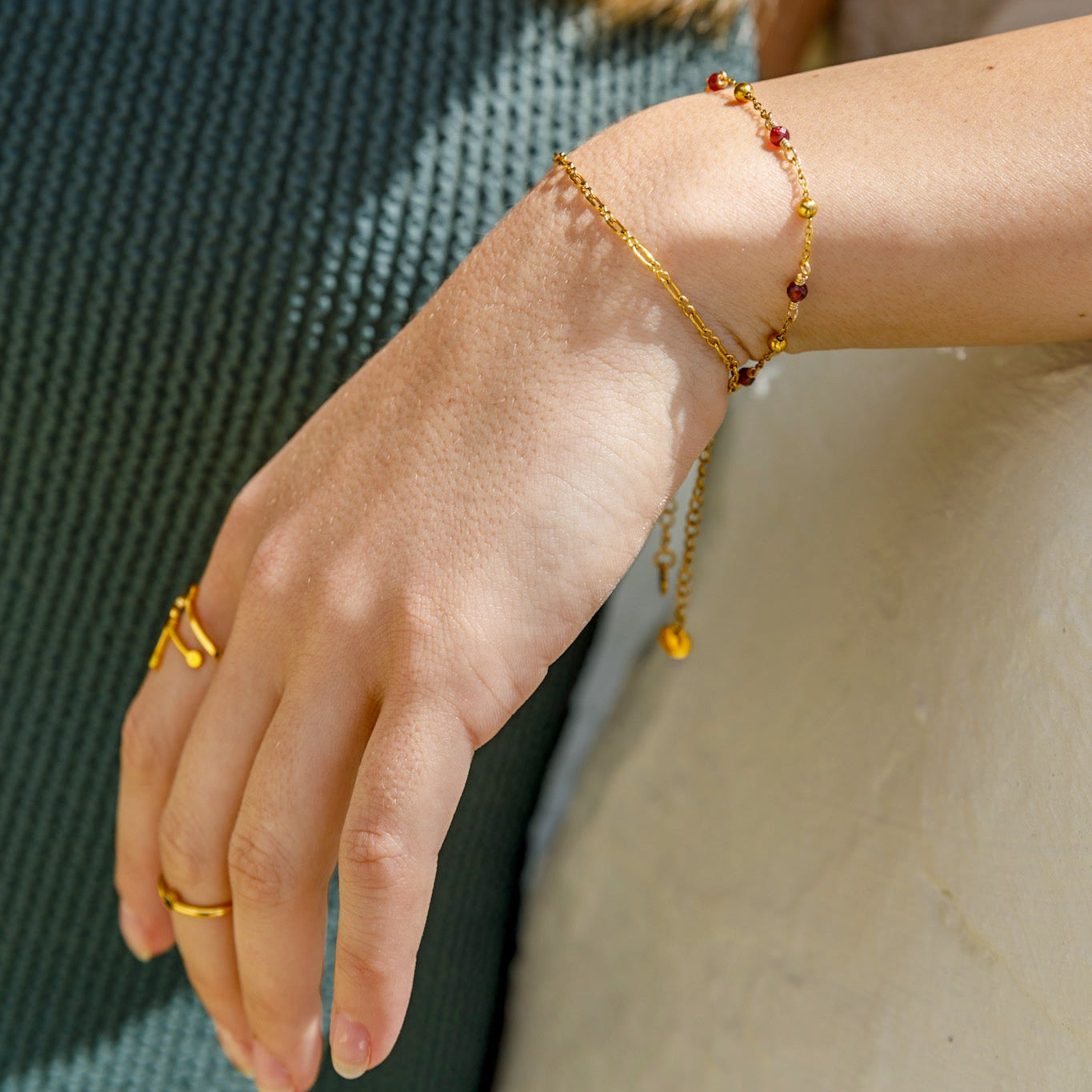 A close-up of a woman's wrist wearing delicate gold chain bracelets and rings, with a textured teal top and a light-colored surface in the background.