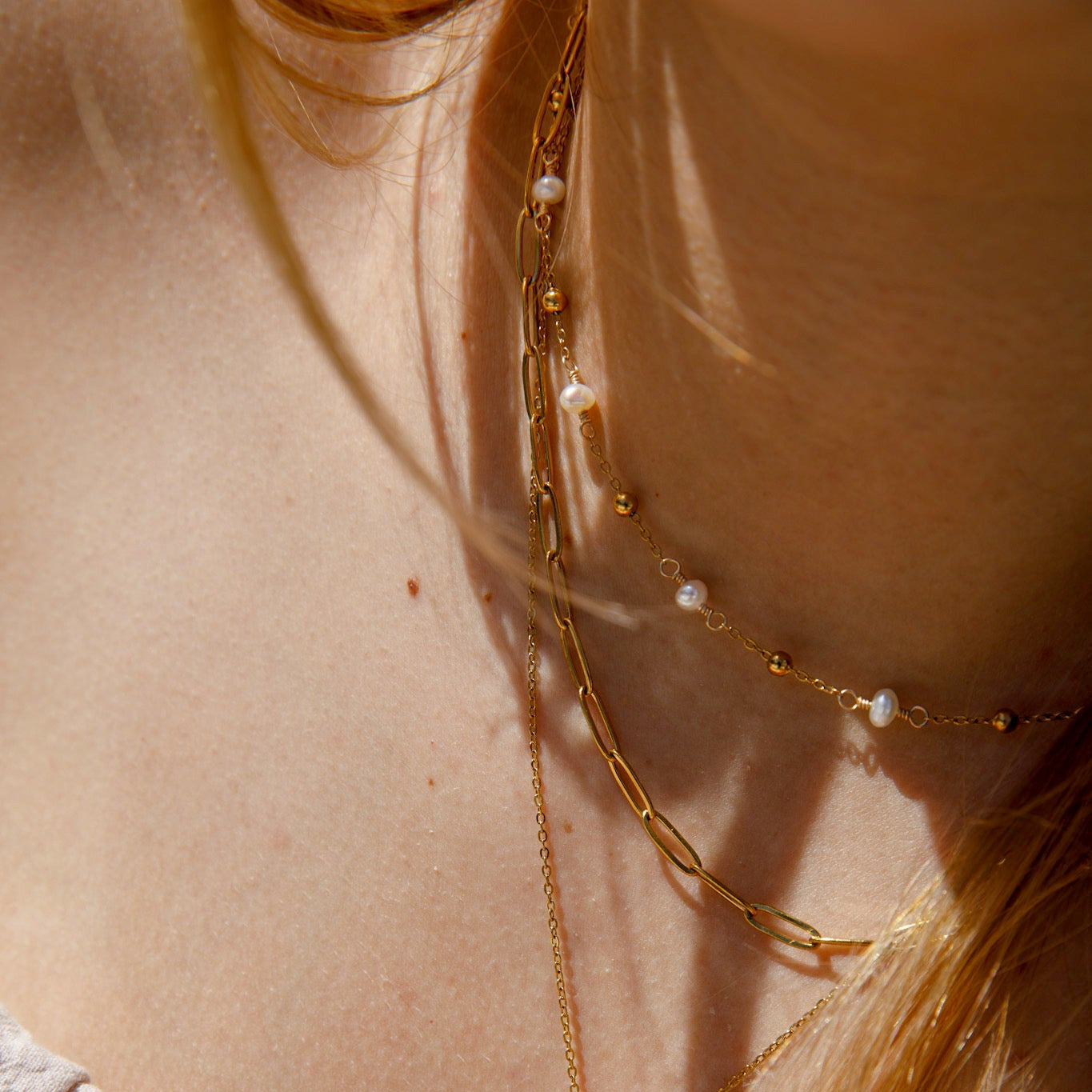 A close-up lifestyle shot of a model's neck and chest wearing a layered gold necklace set. The set includes a gold paperclip chain, a delicate gold chain with small gold beads, and a gold chain with white freshwater pearls.