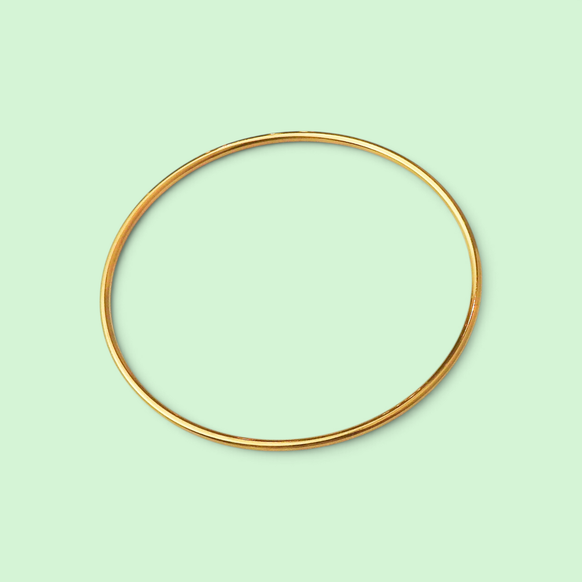 A thin, polished gold bangle bracelet shown against a solid pale mint green background.