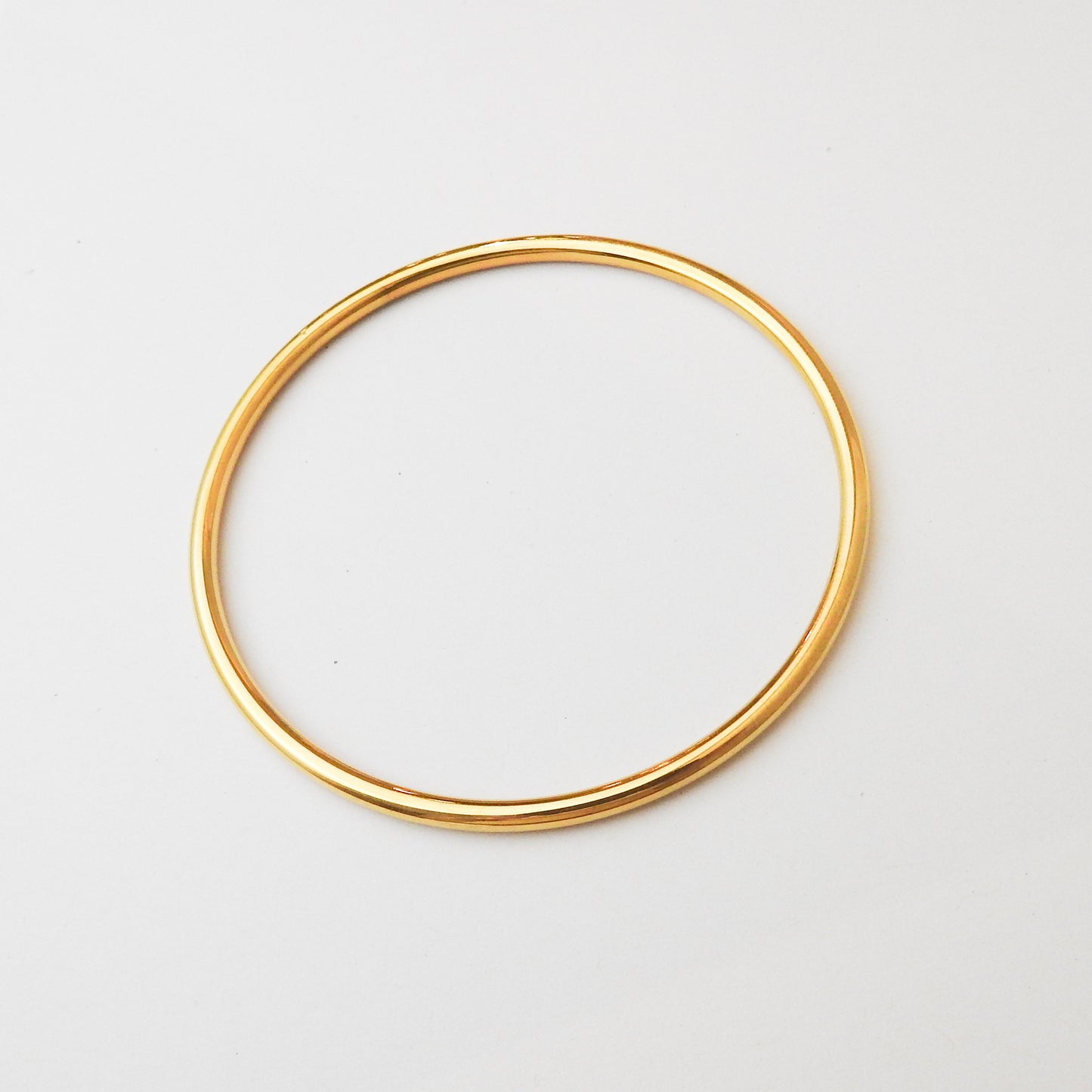A thin, polished gold bangle bracelet shown on a clean, light off-white background.