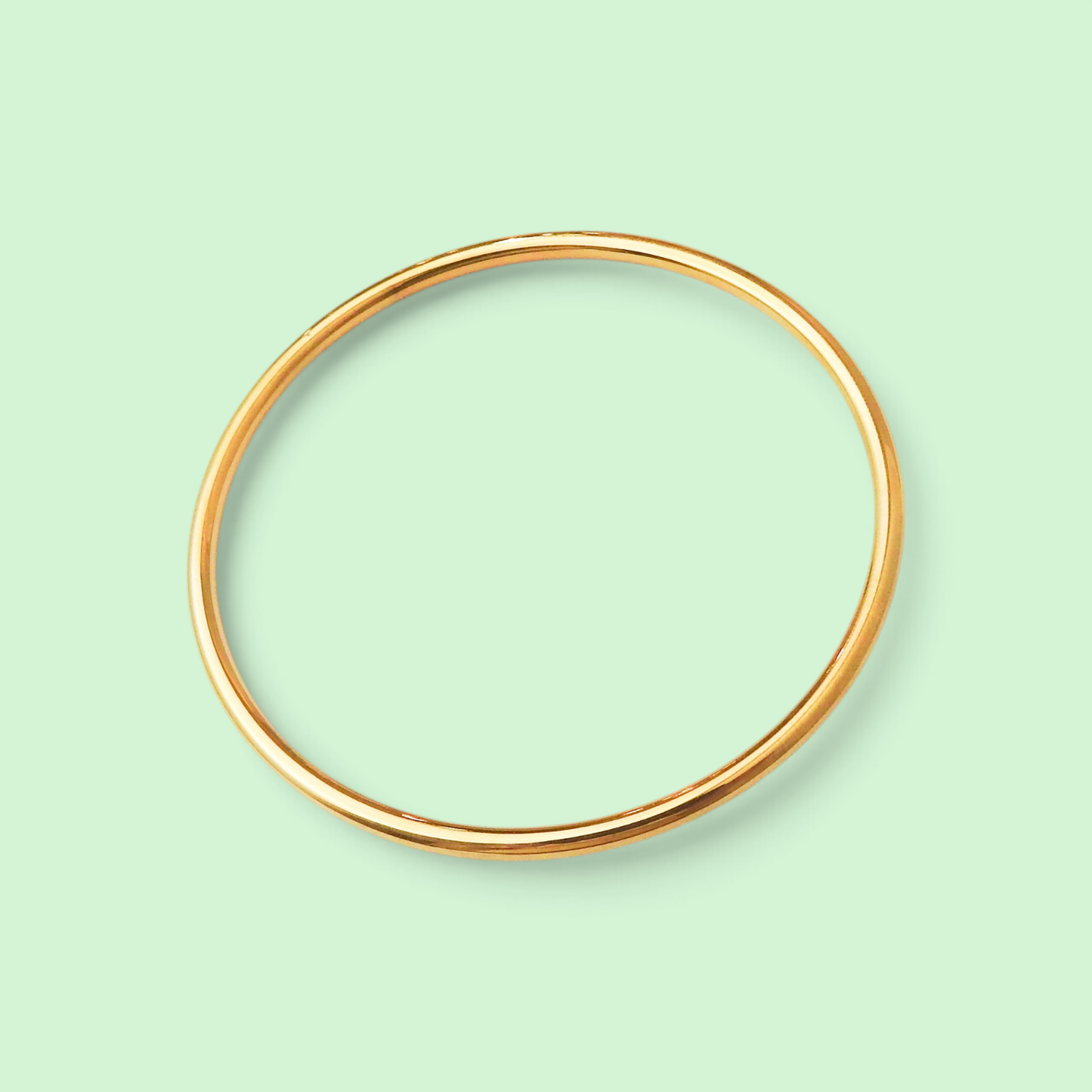 A thin, polished gold bangle bracelet shown against a solid pale mint green background.