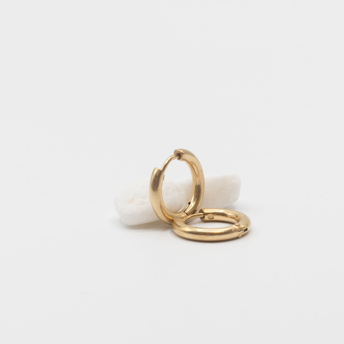 A pair of polished gold huggie hoop earrings resting on a small white stone against a clean white background.