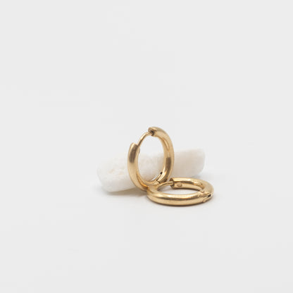 A pair of polished gold huggie hoop earrings resting on a small white stone against a clean white background.