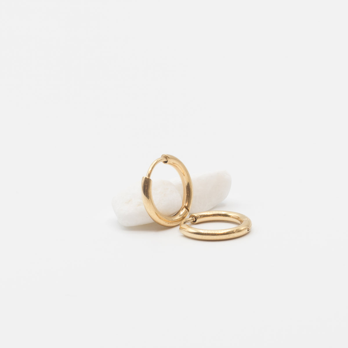 A pair of gold huggie hoop earrings displayed on a small white stone against a clean, light gray background.