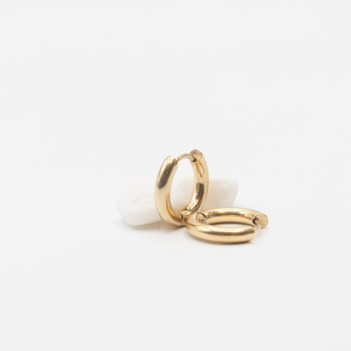 Two small gold huggie hoop earrings resting on a smooth white stone against a clean white background.