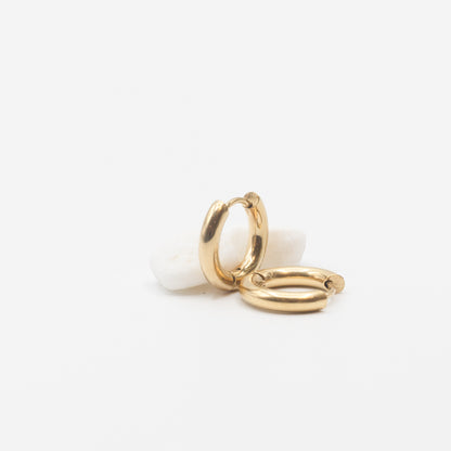 Two small gold huggie hoop earrings resting on a smooth white stone against a clean white background.