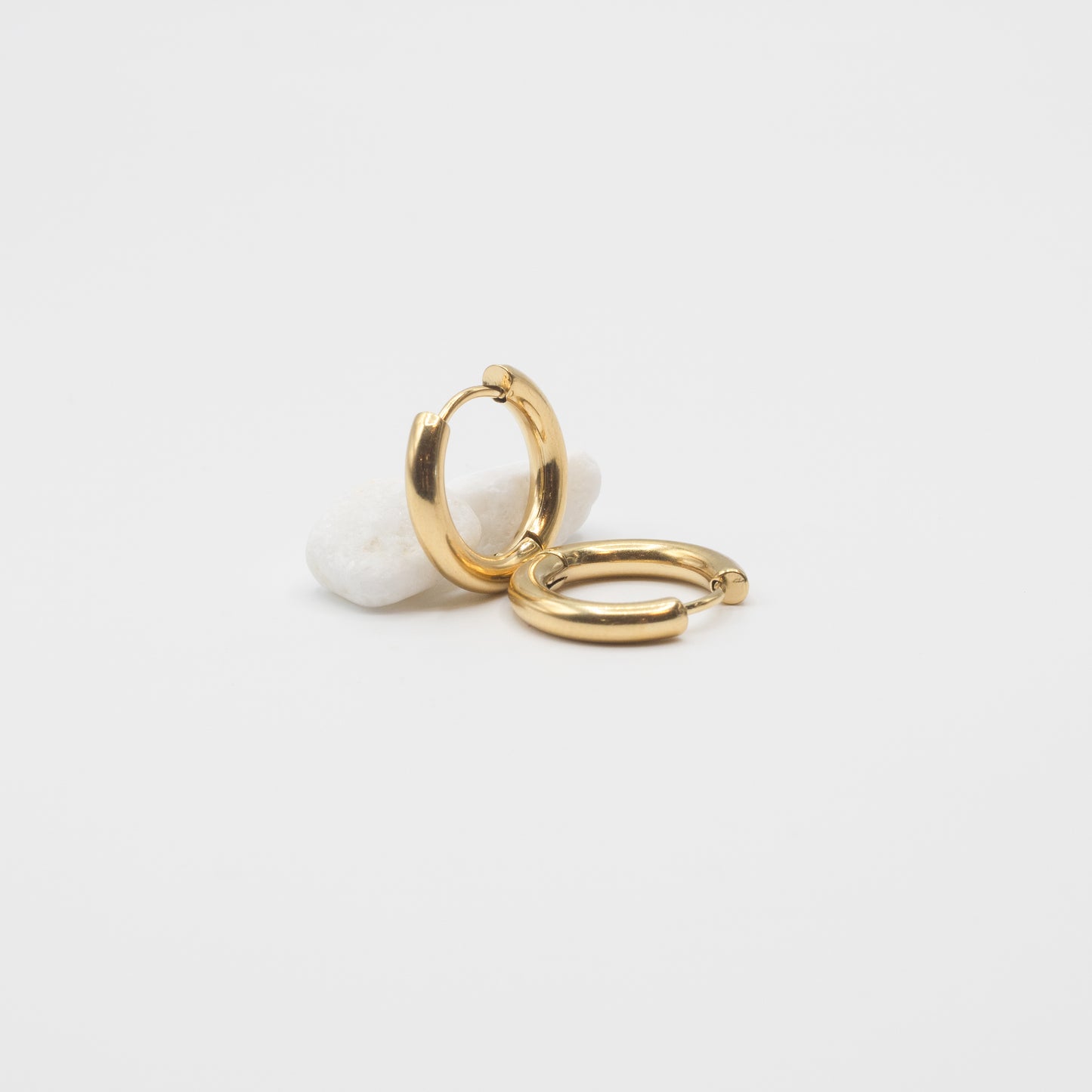 Two polished gold huggie hoop earrings displayed on a small white stone against a clean white background.