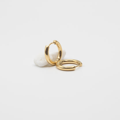Two polished gold huggie hoop earrings displayed on a small white stone against a clean white background.