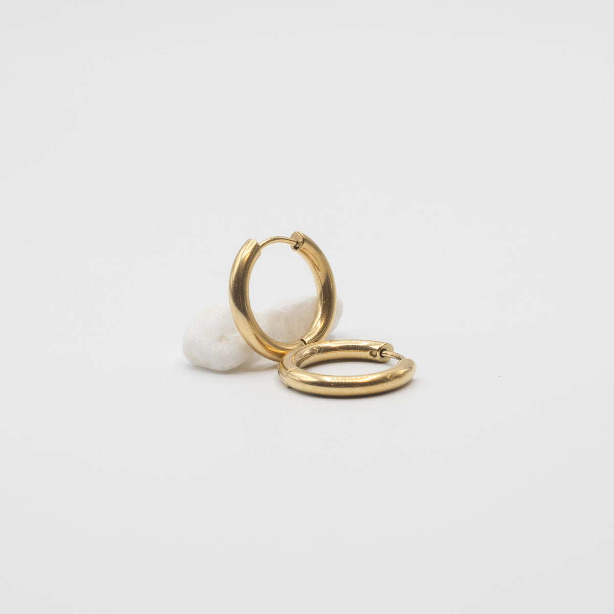 A pair of polished gold huggie hoop earrings resting on a small white textured stone against a clean, light gray background.