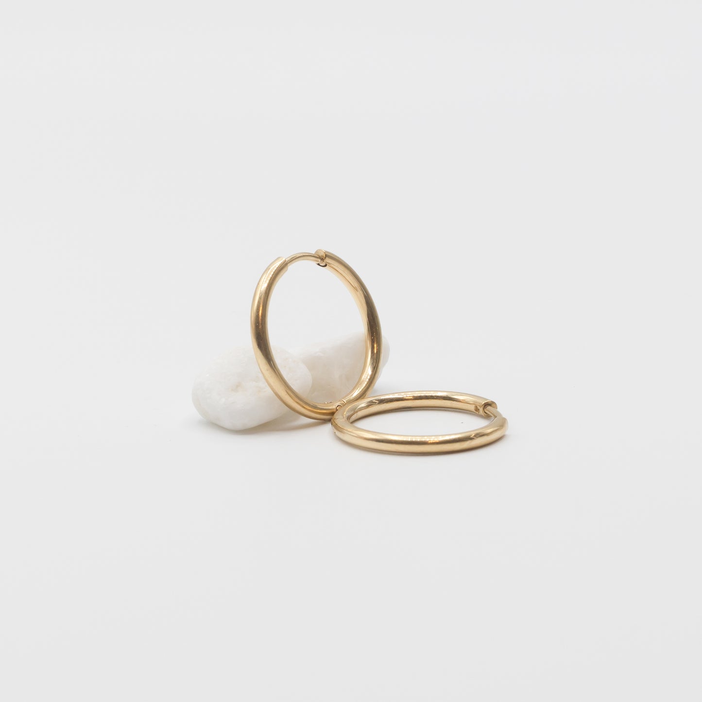 Two thin gold hoop earrings resting on a small white stone against a clean white background.