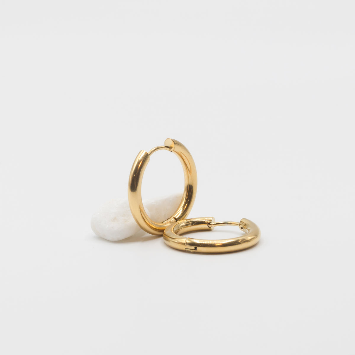 Two polished gold huggie hoop earrings resting on a small white stone against a clean white background.