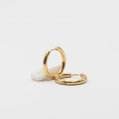 Two polished gold huggie hoop earrings resting on a small white stone against a clean white background.