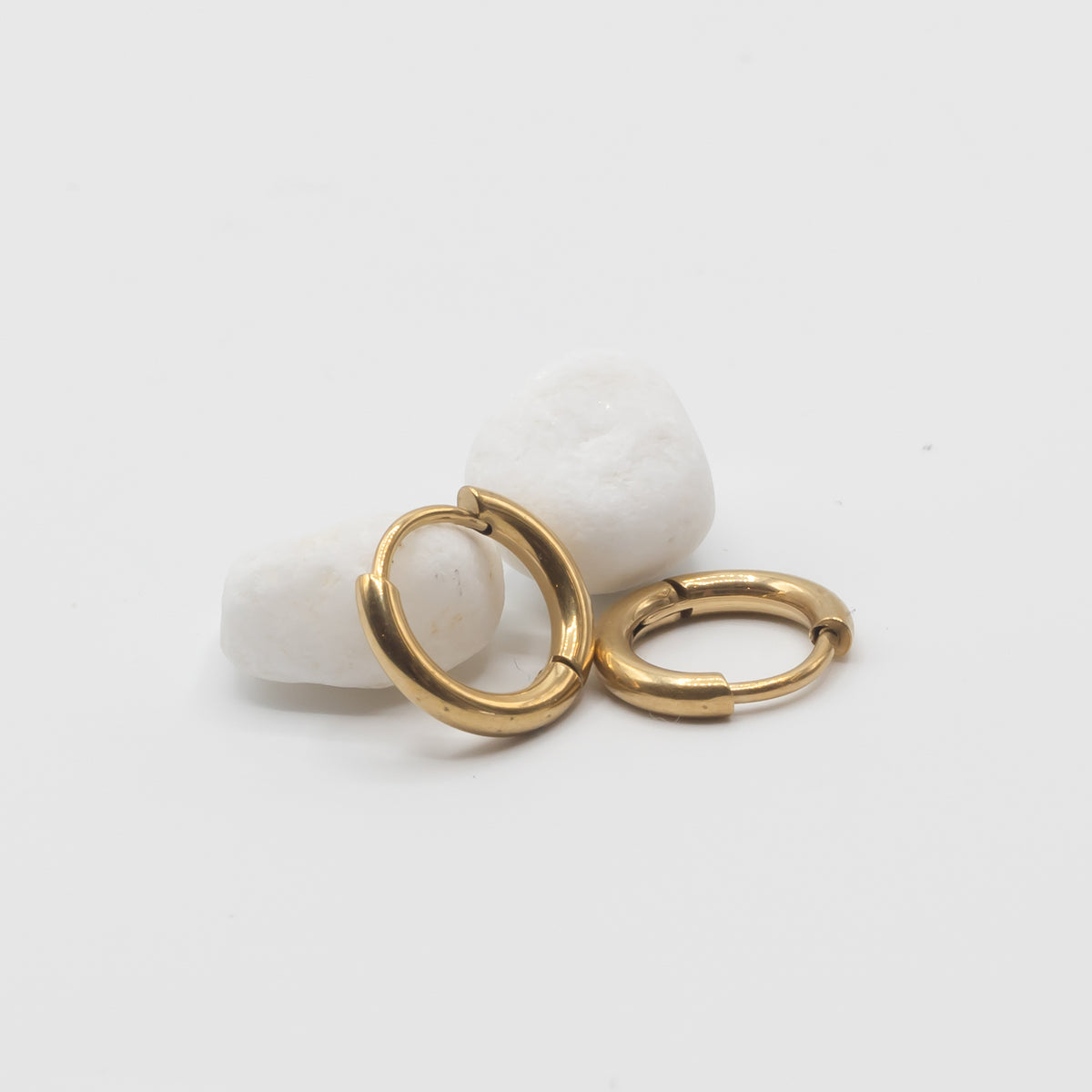 Two small gold hoop earrings resting on two smooth white stones against a clean white background.