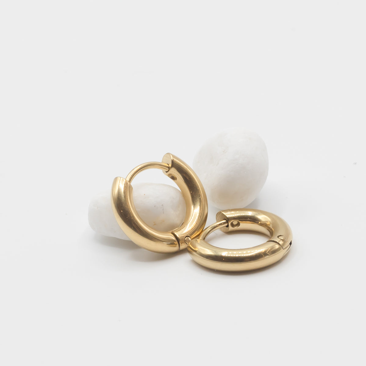 A pair of gold huggie hoop earrings displayed on small white stones against a clean white background.