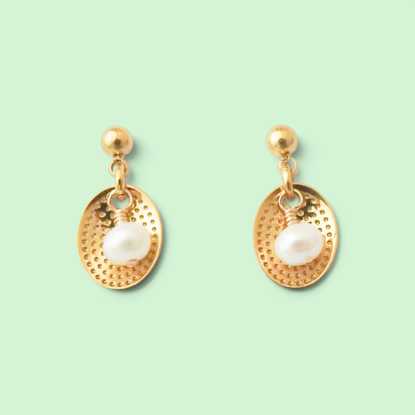 A pair of gold dangle earrings with textured oval charms, each featuring a white freshwater pearl, displayed on a solid pale mint green background.