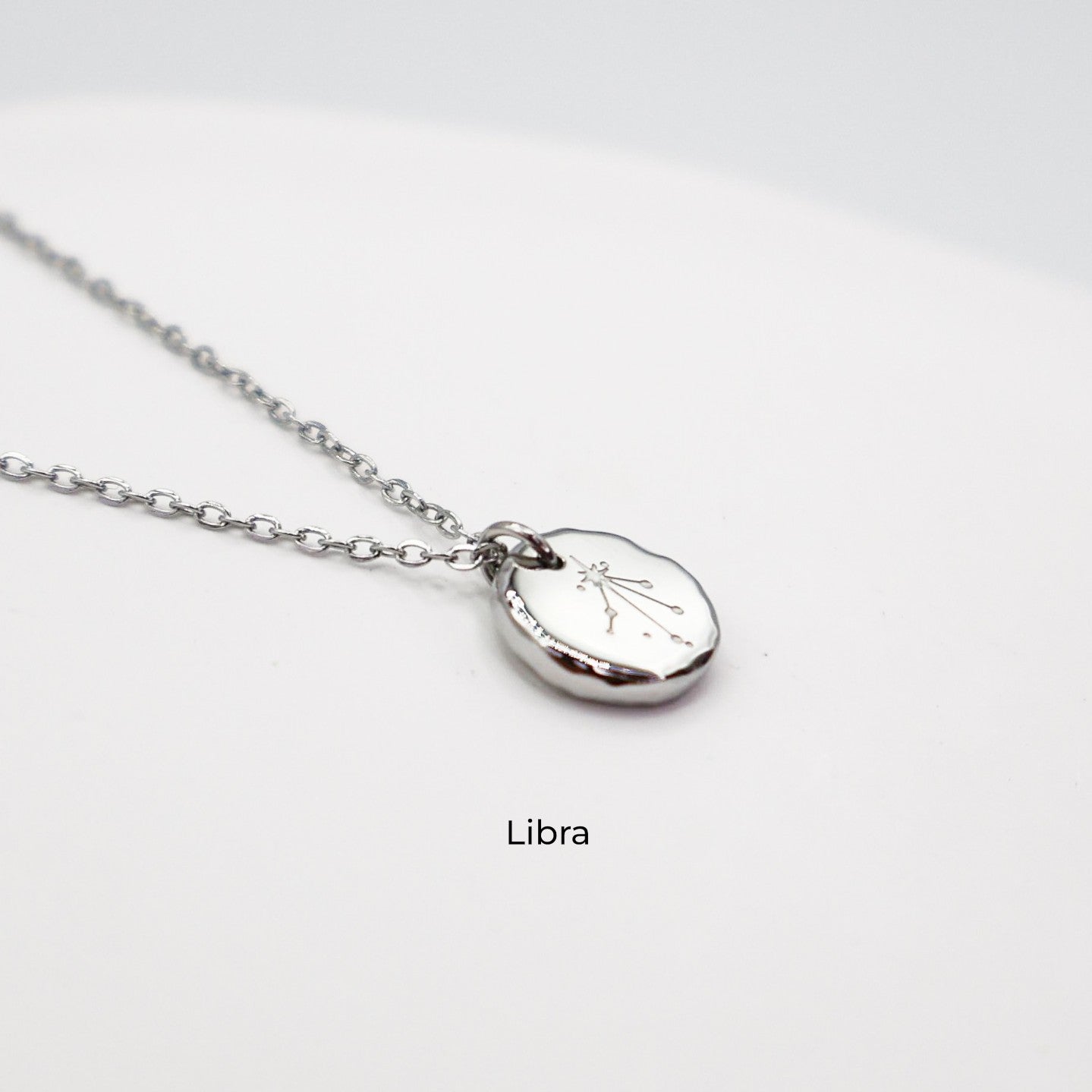 Silver necklace with a round pendant on a light gray background. The pendant is engraved with the Libra star sign constellation