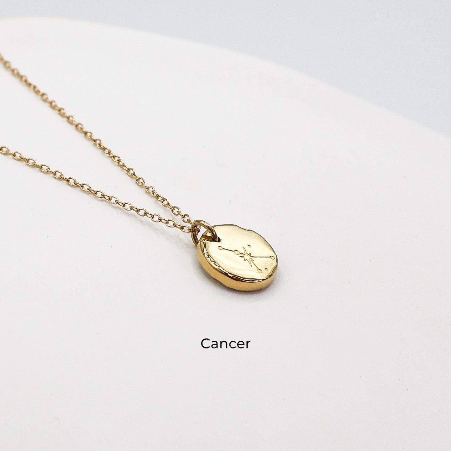 Gold necklace with a round pendant on a light gray background. The pendant is engraved with the Cancer star sign constellation