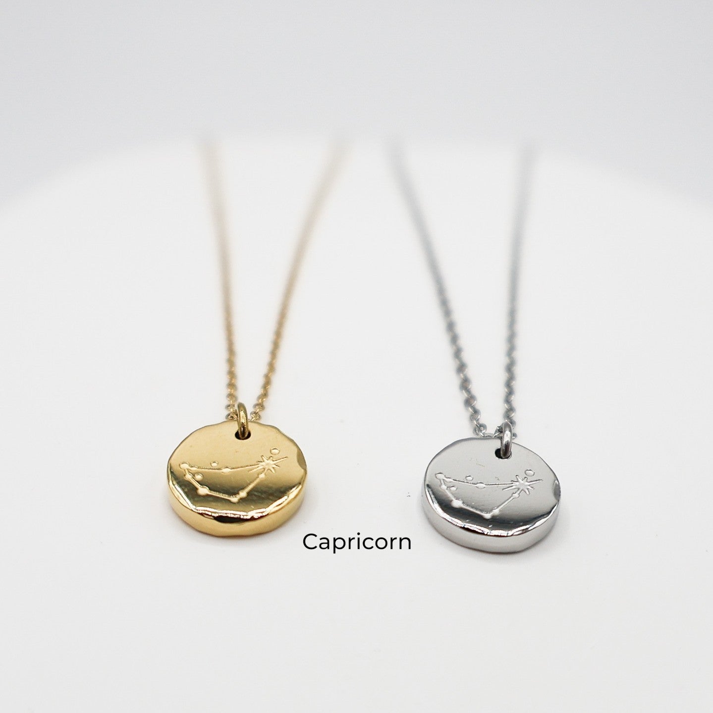 Two necklaces with zodiac symbols, one gold and one silver, on a white background. The necklaces are engraved with the Capricorn star sign constellation