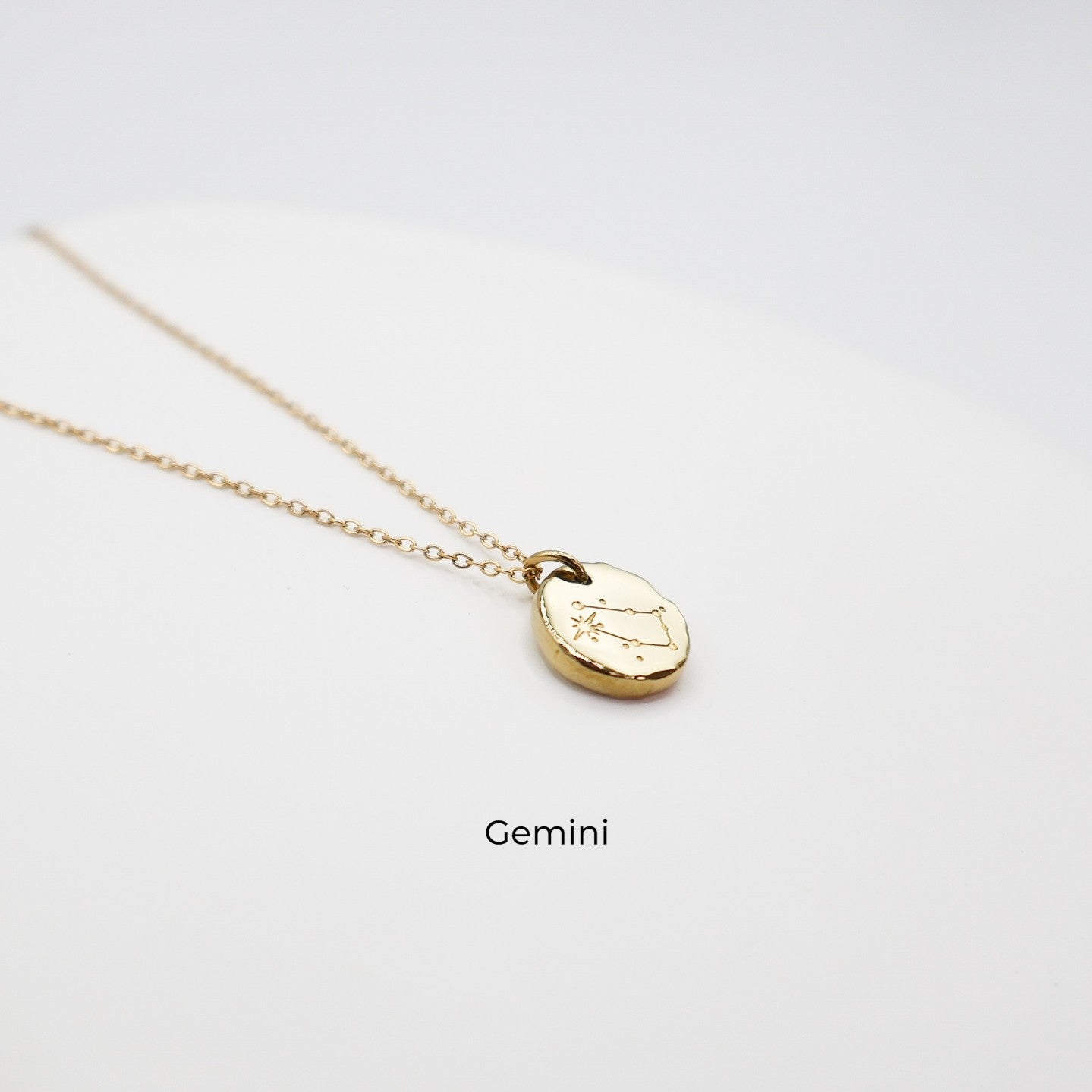 Gold necklace with a round pendant on a light gray background. The pendant is engraved with the Gemini star sign constellation