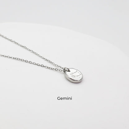 Silver necklace with a round pendant on a light gray background. The pendant is engraved with the Gemini star sign constellation