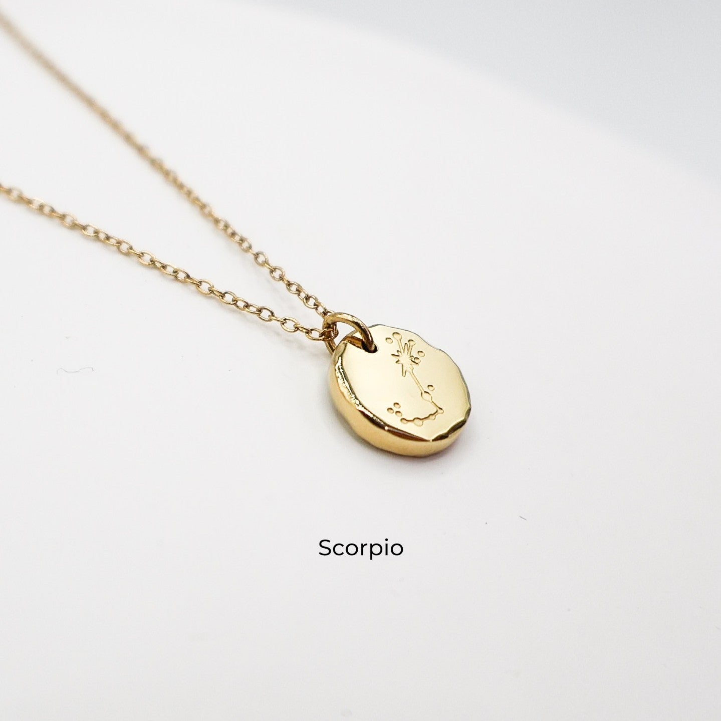 Gold necklace with a round pendant on a light gray background. The pendant is engraved with the Scorpio star sign constellation