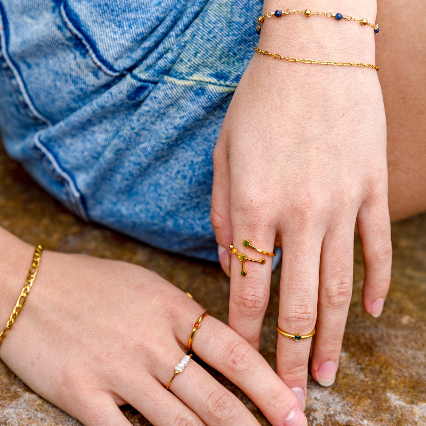 A close-up lifestyle shot of a person's hands wearing multiple delicate gold rings and bracelets, with blue denim shorts visible in the background.