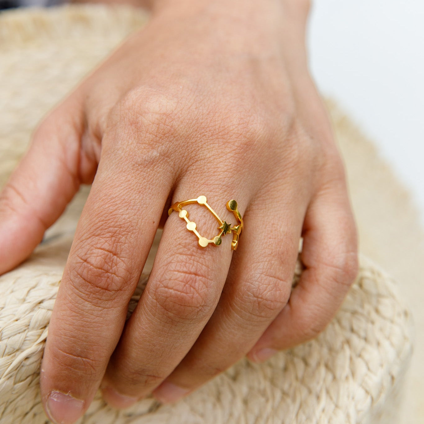 A gold ring with a wavy, openwork design and small green leaf accents, shown worn on a person's finger resting on a woven straw surface.