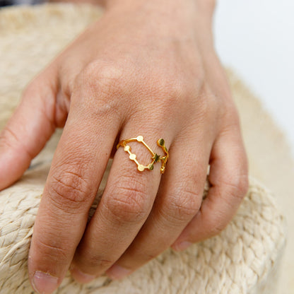 A gold ring with a wavy, openwork design and small green leaf accents, shown worn on a person's finger resting on a woven straw surface.