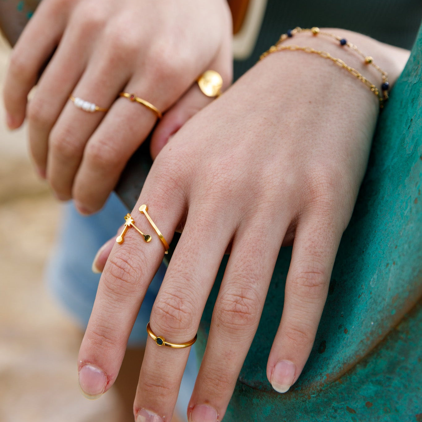 Close-up of a person's hands wearing multiple delicate gold rings and bracelets, with a teal top and sunglasses in the background.