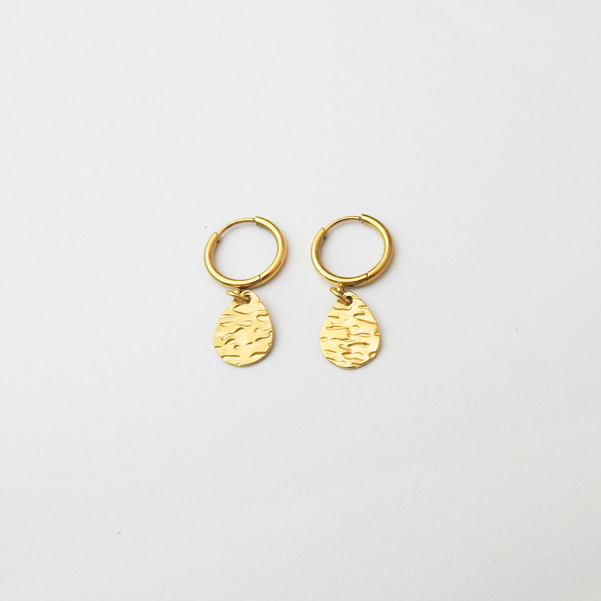 A pair of small gold huggie hoop earrings with textured teardrop charms, displayed on a clean white background.