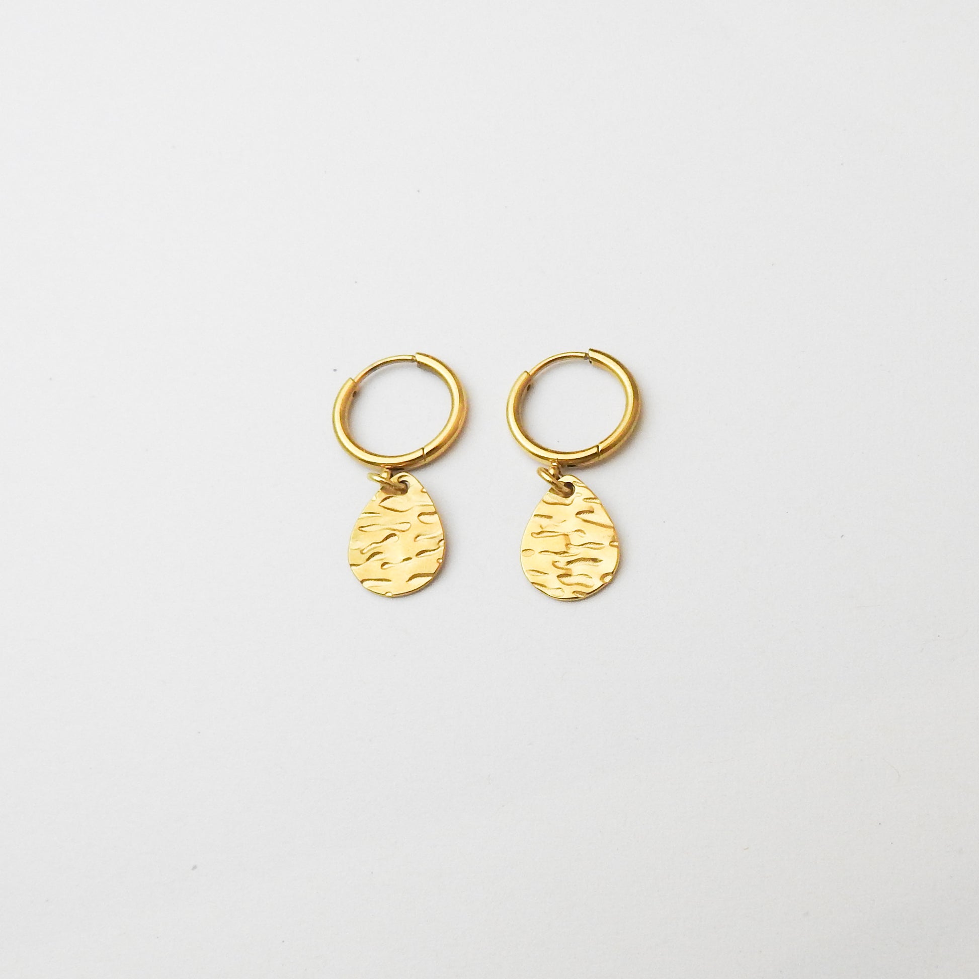 A pair of small gold huggie hoop earrings with textured teardrop charms, displayed on a clean white background.