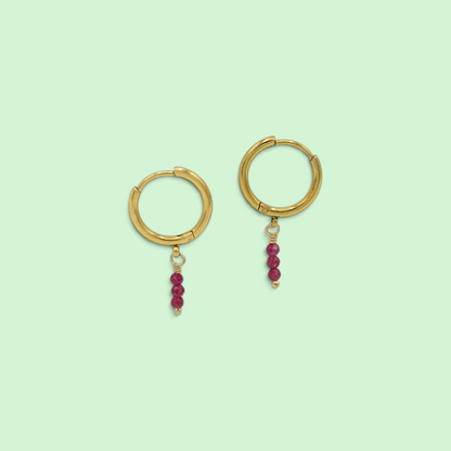 A pair of gold hoop earrings with small dangling clusters of dark pinkish-red beads, shown on a solid pale mint green background.