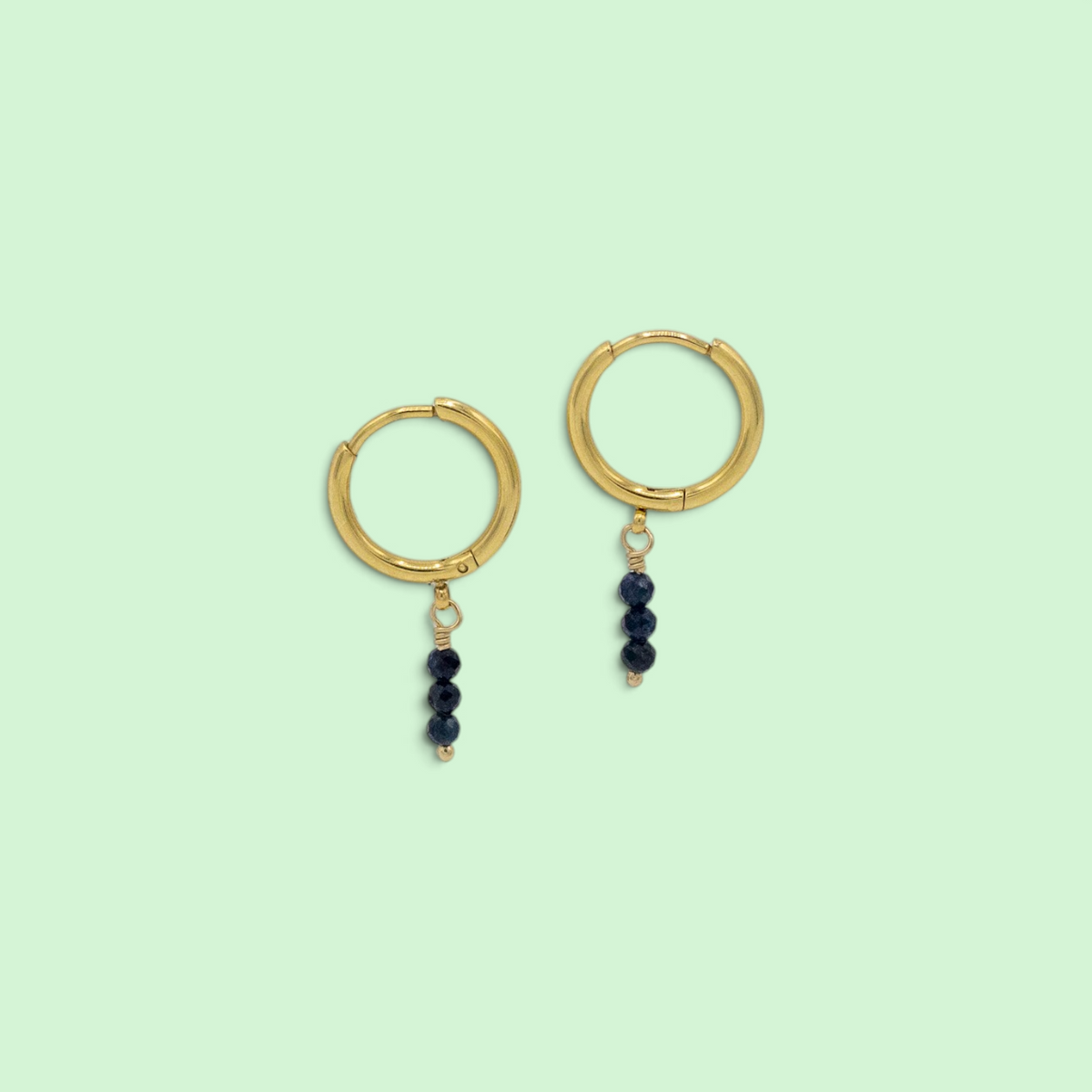 A pair of gold hoop earrings with three small dark beads dangling from each hoop, shown on a solid pale mint green background.