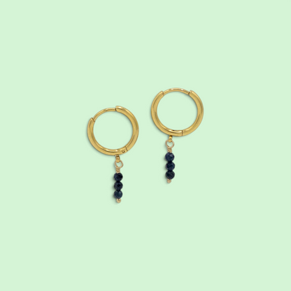 A pair of gold hoop earrings with three small dark beads dangling from each hoop, shown on a solid pale mint green background.