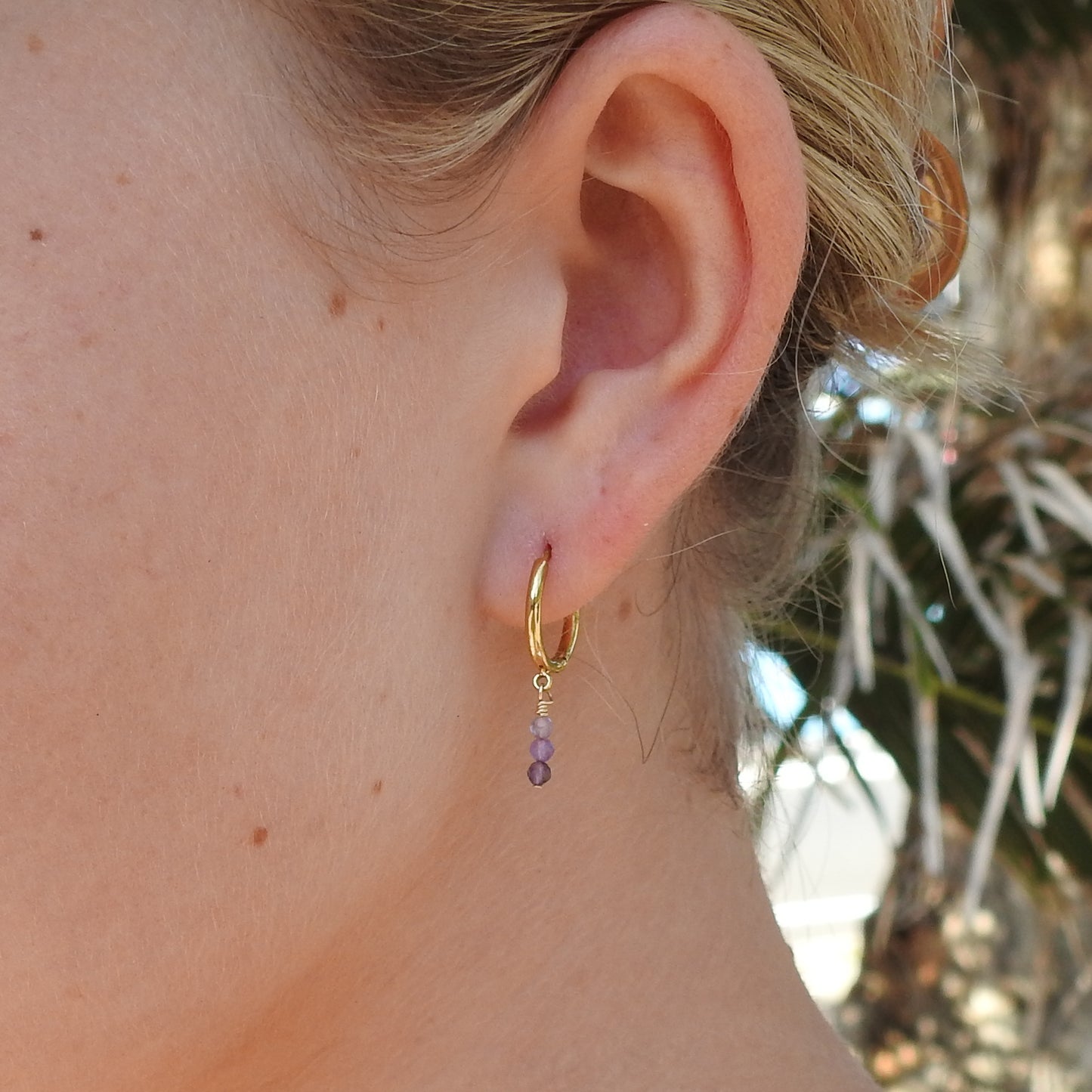 A close-up of a person's ear wearing a small gold hoop earring with a dangling purple bead charm, set against a softly blurred outdoor background.