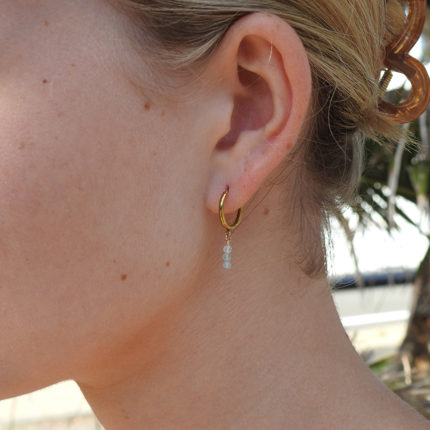A close-up of a person's ear wearing a small gold hoop earring with a dangling clear bead, photographed outdoors with blurred foliage in the background.