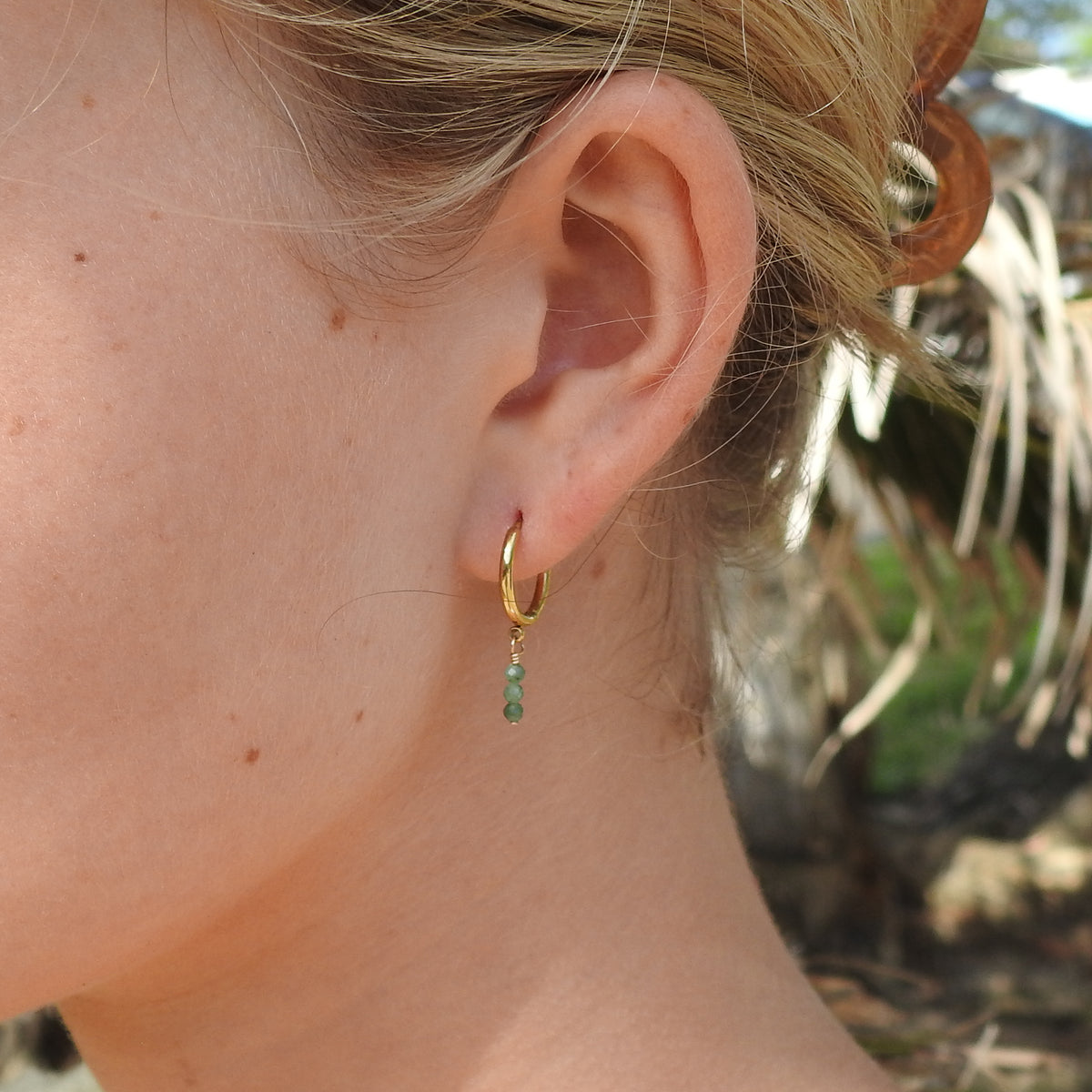 A close-up side profile of a woman's ear wearing a small gold hoop earring with a dangling green bead, set against a blurred outdoor background.