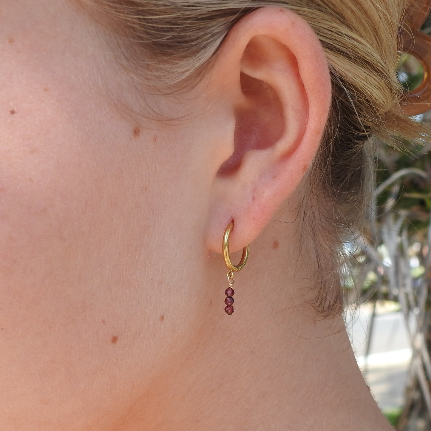 A close-up of a person's ear wearing a small gold hoop earring with three dangling faceted burgundy beads.