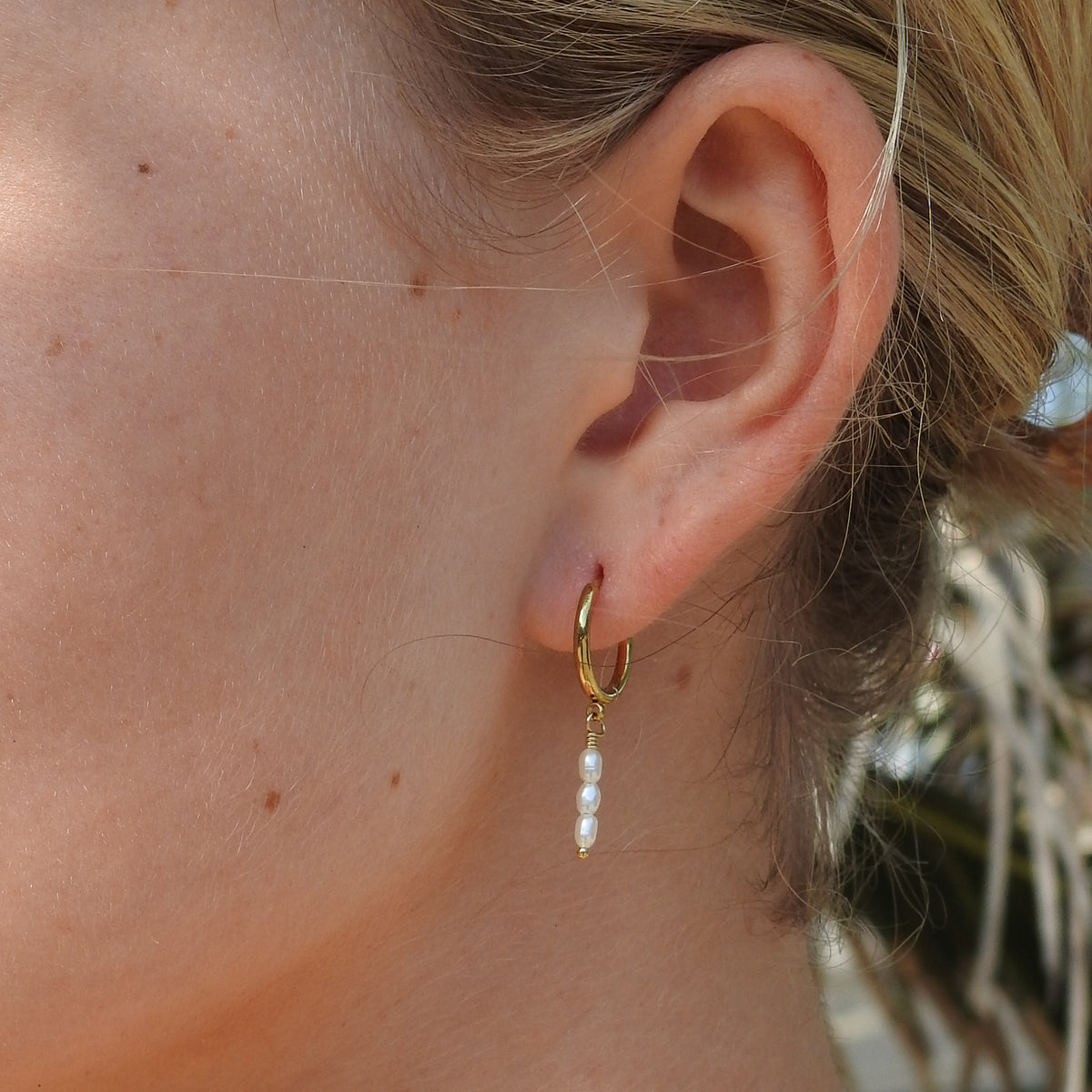 A close-up of a person's ear wearing a small gold hoop earring with a dangling pearl bead drop.