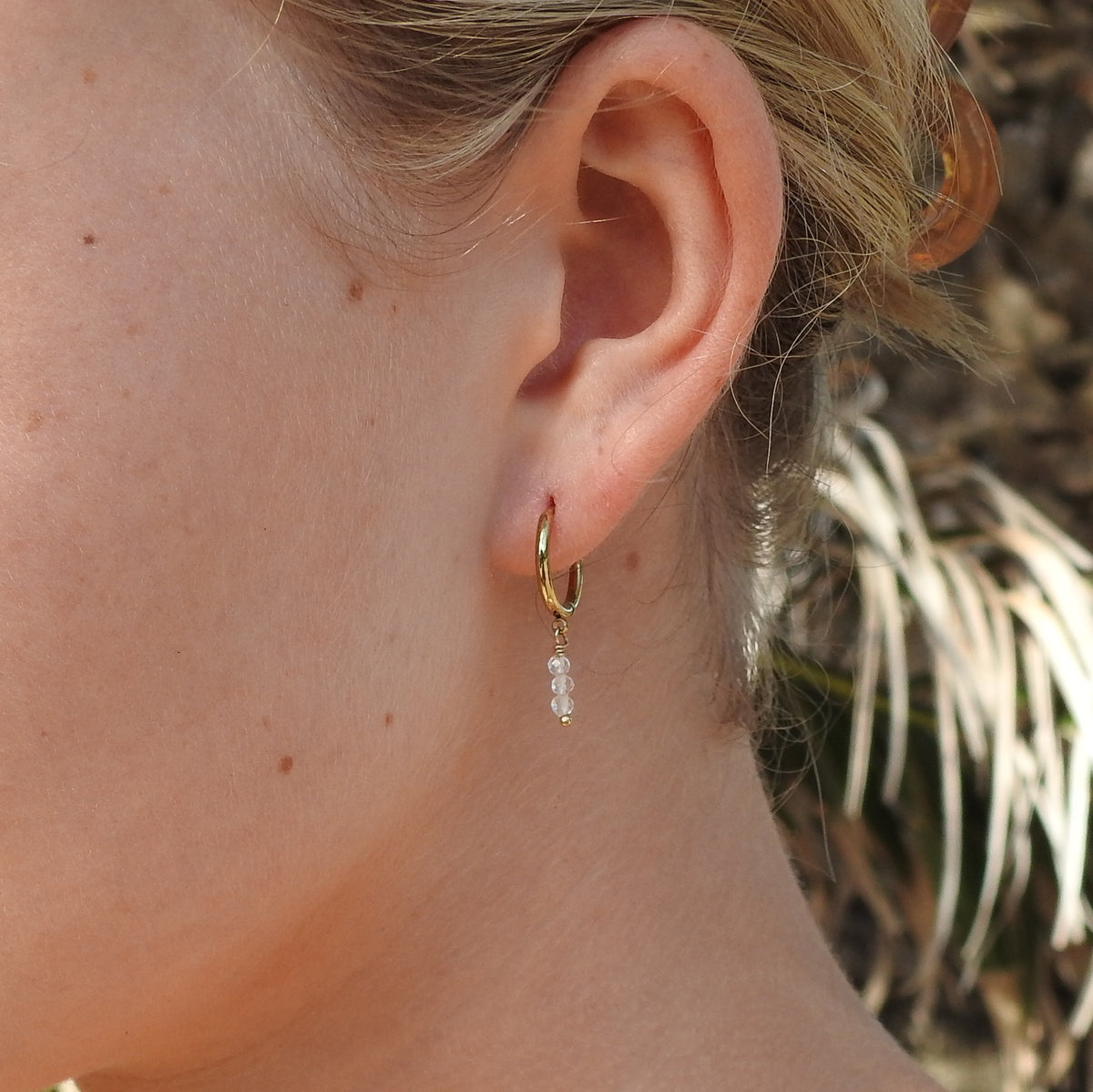A close-up of a person's ear wearing a small gold hoop earring with a dangling clear faceted bead charm, against a softly blurred outdoor background.