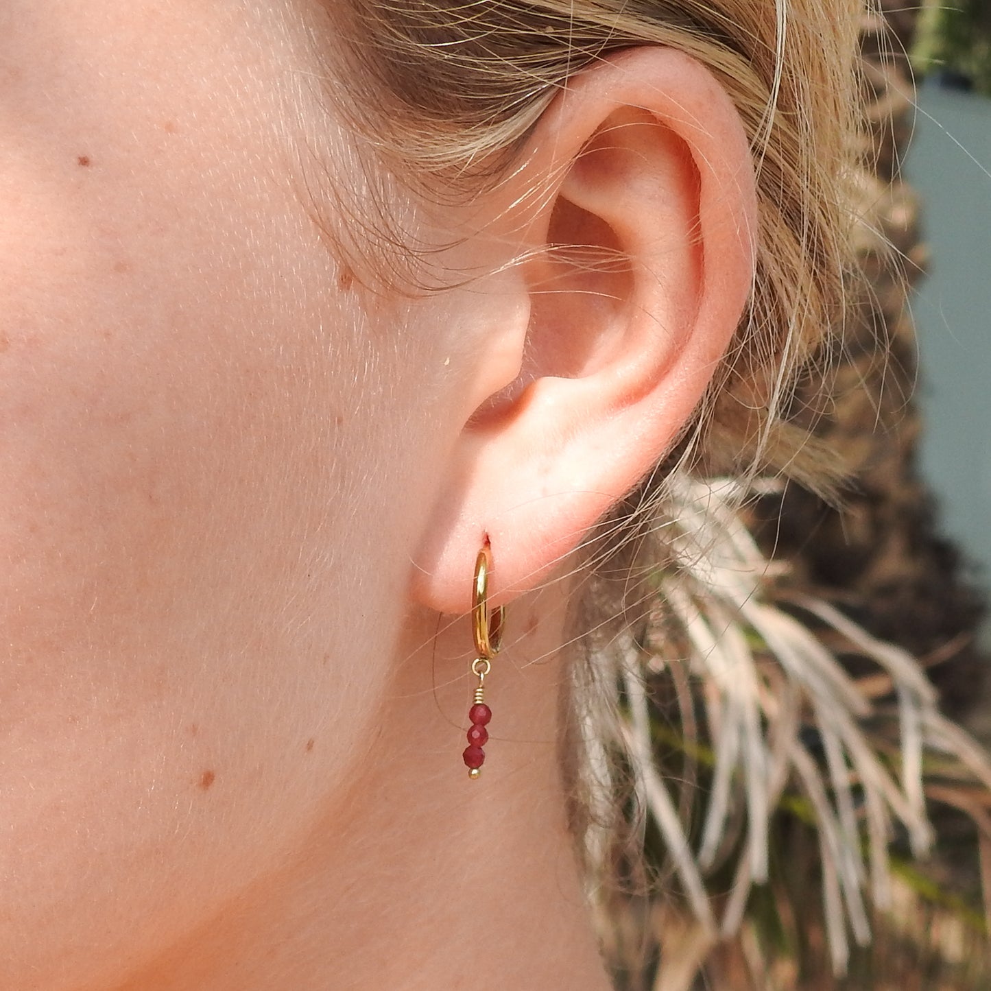 A close-up of a person's ear wearing a small gold hoop earring with a dangling chain and three small red beads.